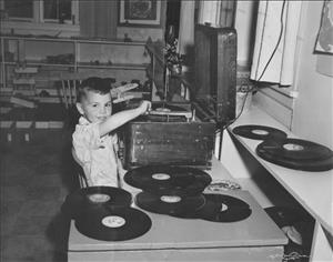 A young white boy smiles while he lowers the needle on a record on a large wooden record player. The table in front of him is strewn with records